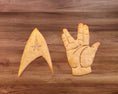 Load image into Gallery viewer, Two cookie shapes on a wooden surface: one resembling a starship and the other a hand making a gesture.