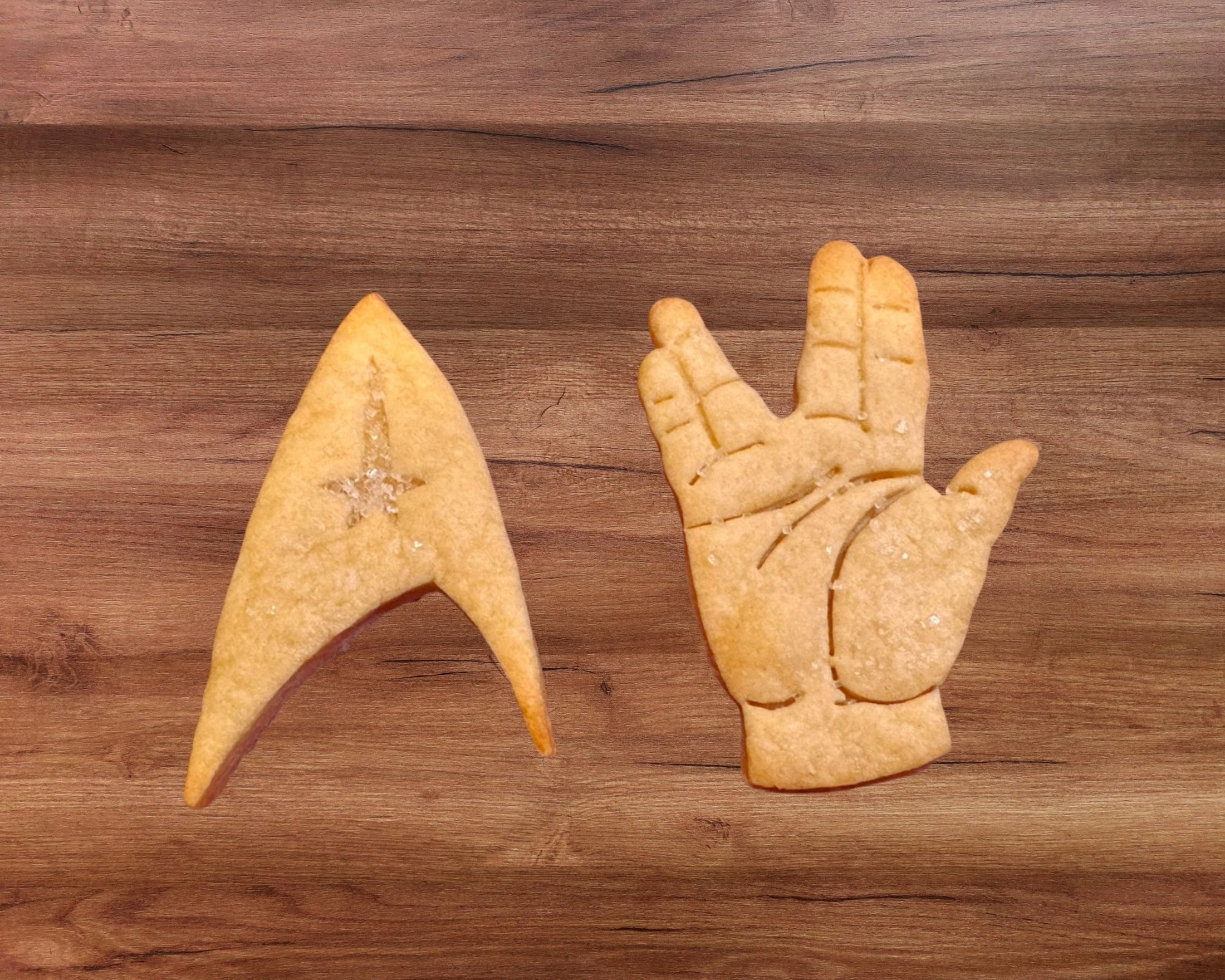 Two cookie shapes on a wooden surface: one resembling a starship and the other a hand making a gesture.